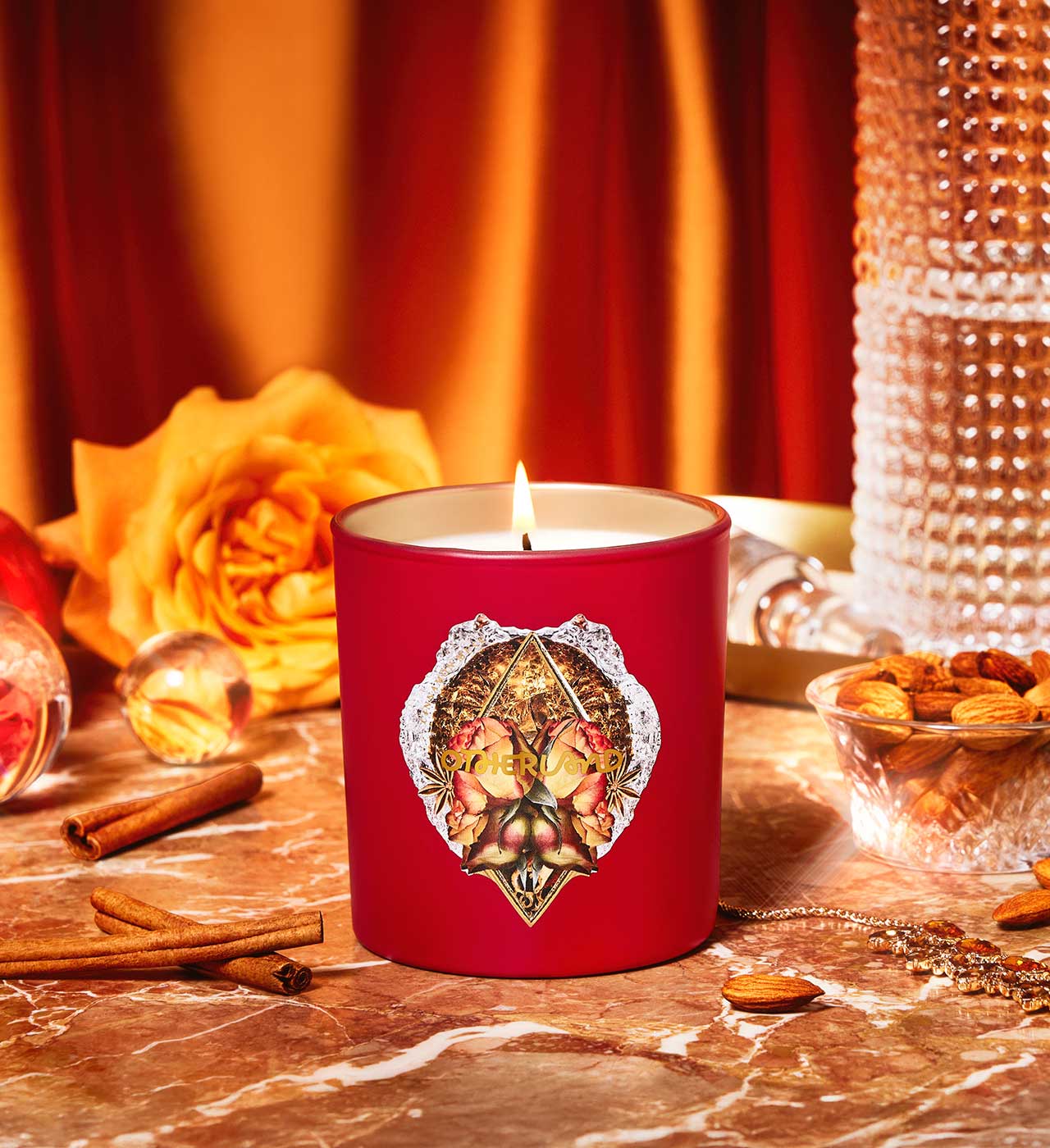 Red candle with decorative lid on a table with cinnamon sticks, rose, and nuts.
