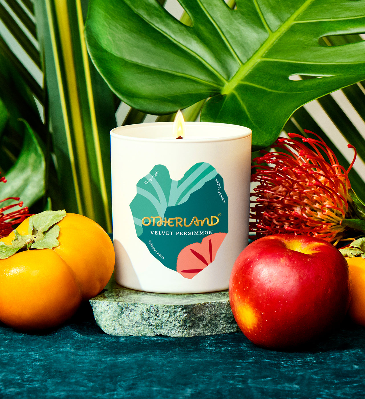 Candle with 'Otherland' branding surrounded by fruits and leaves